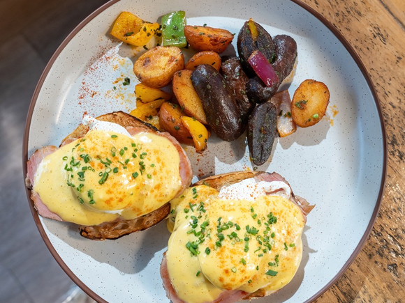 A plate of breakfast eggs benedict with two poached eggs on ham, plus roasted potatoes and vegetables.