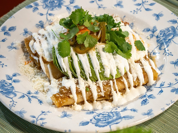 A plated dish with breaded cutlets topped with white sauce, sliced cucumbers, olives, and chopped cilantro.