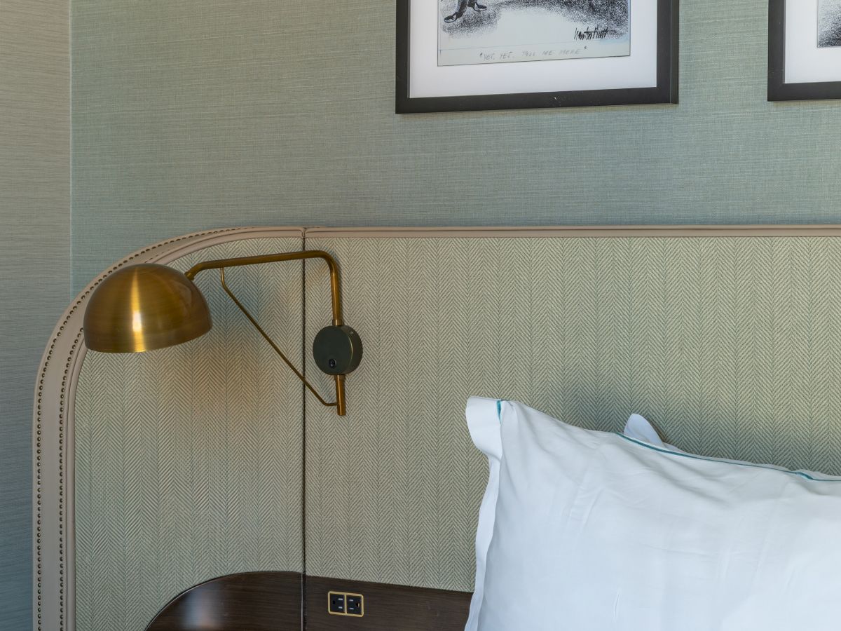 A hotel bedroom scene with a bed, brass reading lamp, and framed wall art above the headboard.