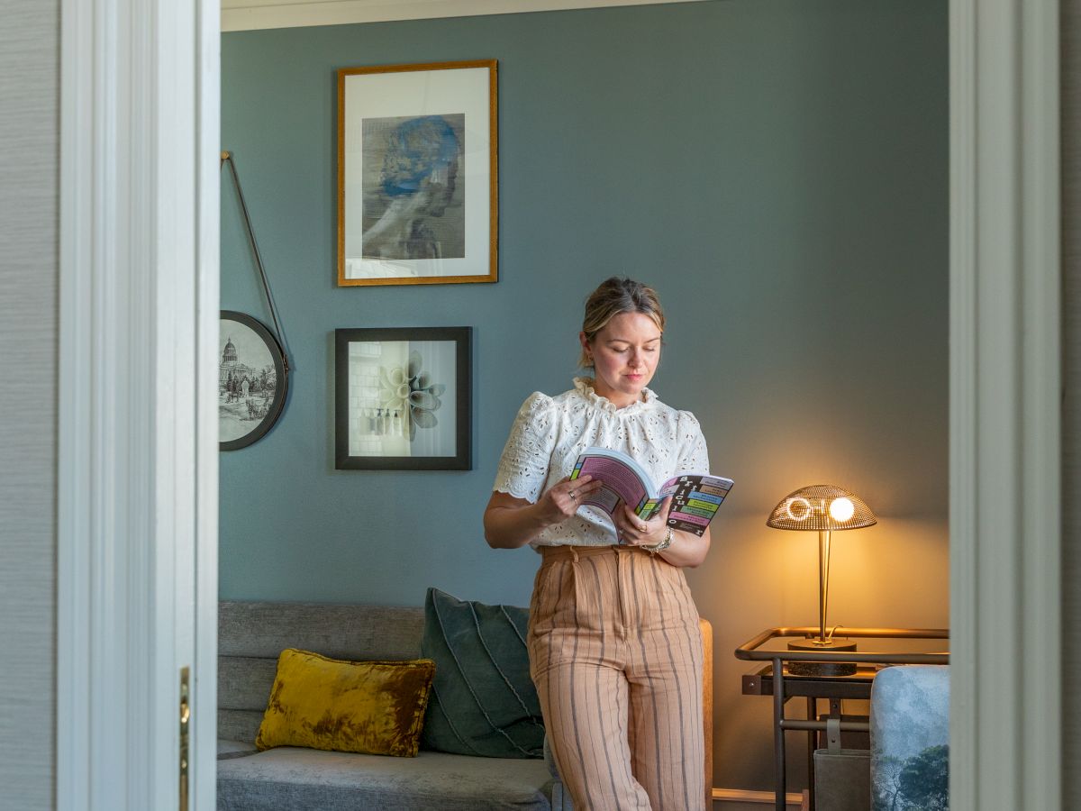 A woman stands in a cozy living room reading a magazine by a lamp.