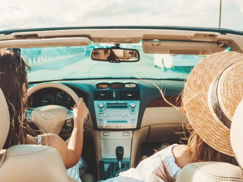 Inside a car, two people ride in the front seats while driving down a sunny road.