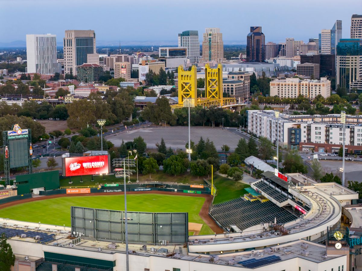 A city skyline with a baseball stadium in the foreground and a bright yellow stadium scoreboard tower in the center, at dusk.