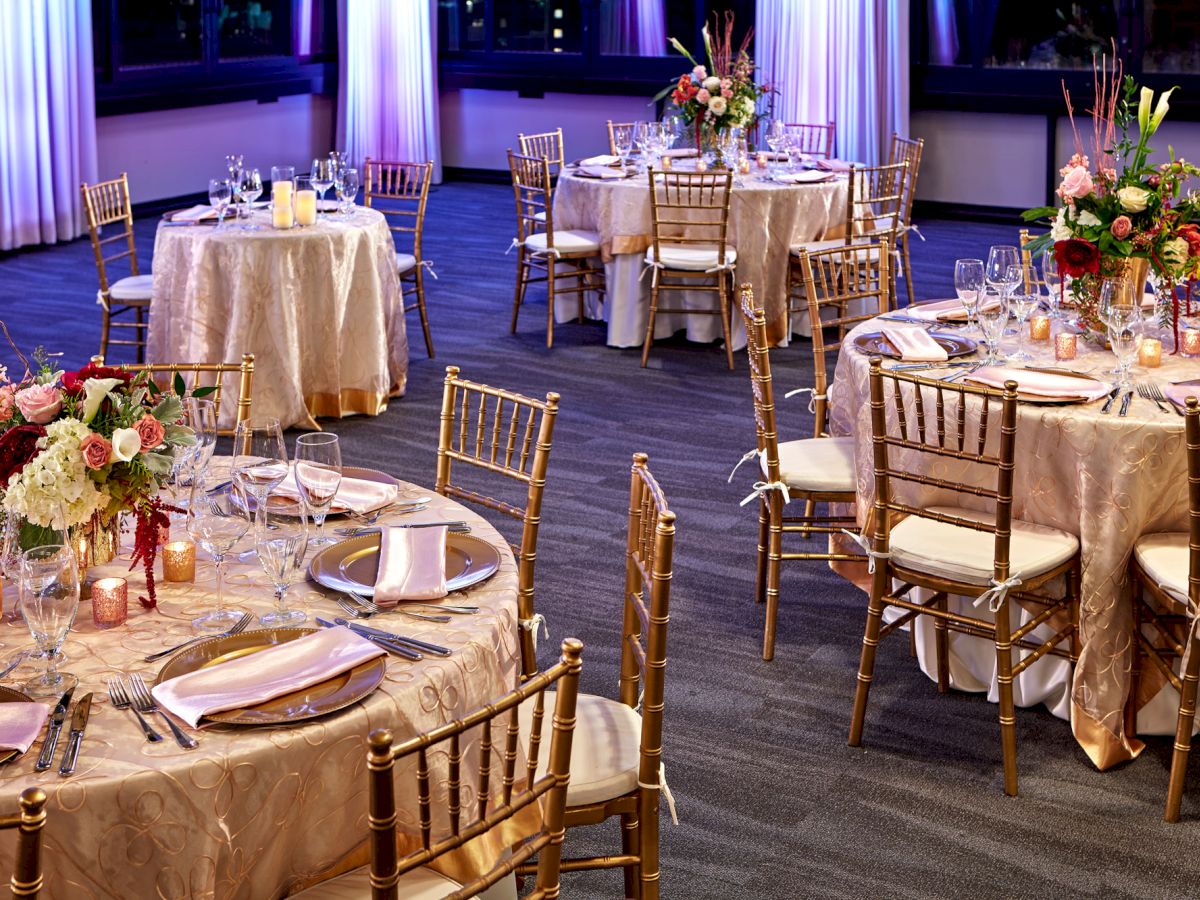 Elegant banquet tables set with gold chairs, white linens, crystal glasses, candle jars, and colorful flower centerpieces.