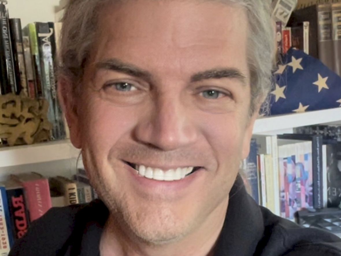 A smiling man with gray hair sits indoors in front of shelves of books and a small American flag cushion.